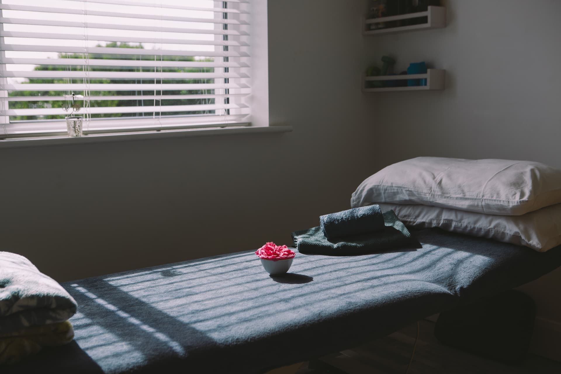 Treatment room with massage table, soft natural light through window blinds, folded towels and a pink flower