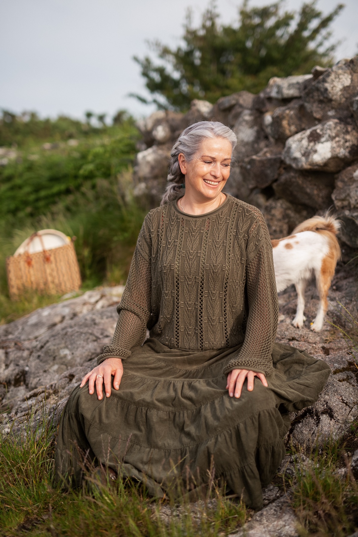 A woman sitting peacefully among rocks in a grassy landscape, a small dog nearby